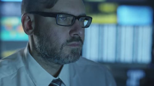 Manager is working on a computer in the evening in a dark office room filled with display screens.