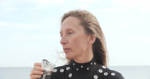 A Young Woman Sits on a Sandy Beach and Drinks Water From a Transparent Plastic Bottle Against the