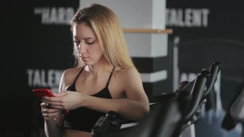 The Girl is Standing in the Gym and Texting on Her Smartphone