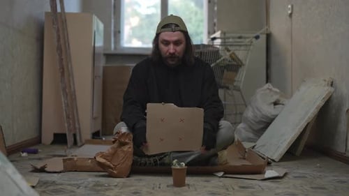 Homeless Poor Man Sitting in a Room of an Abandoned Building with a Money Jar He is Holding a Piece