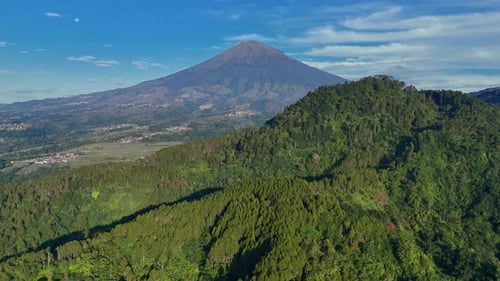 Panoramic aerial drone flight video of green hill with mountain background and cloudy blue sky.
