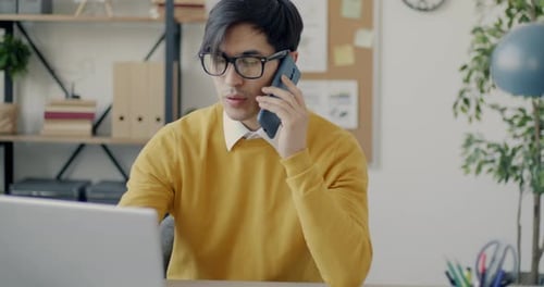 Ambitious Asian Businessman Working with Laptop and Speaking on Mobile Phone in Office