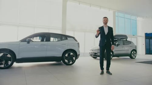 Stylish Caucasian Male in Black Suit Standing in Middle of Car Dealership Showing to Camera Keys to