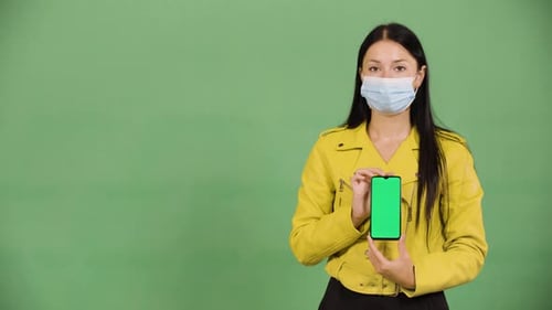 A Caucasian Woman in a Face Mask Shows a Smartphone with Green Screen to the Camera Green Screen