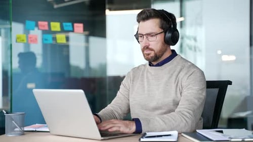 Focused businessman wearing wireless headphones typing on laptop while sitting at desk in office.