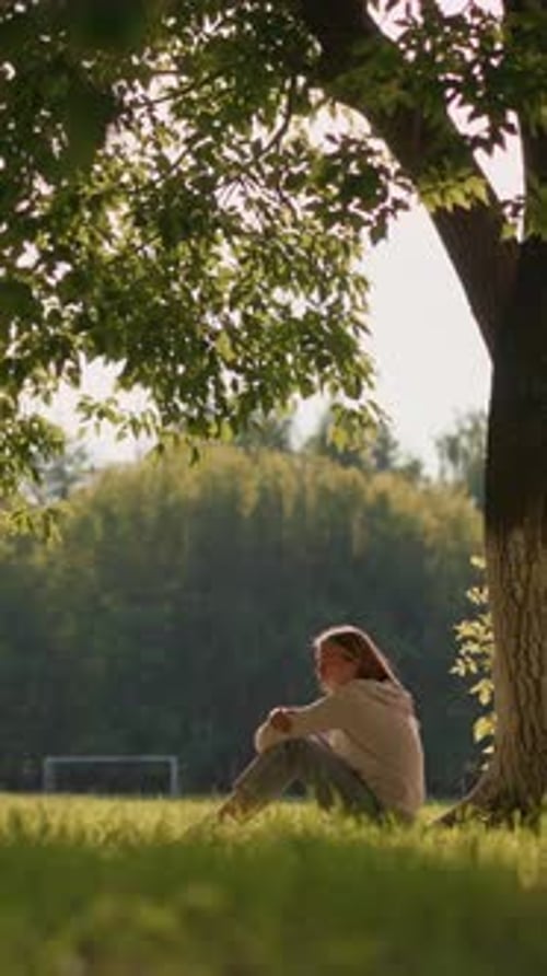 Woman Sitting By Tree in Sunlit Park Surrounded By Greenery and Gentle Breeze
