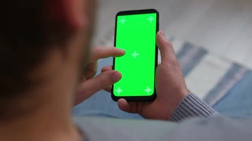 Young Man Sitting at Home Holding Smartphone Green Mockup Screen in Hand Male Person Using Chroma