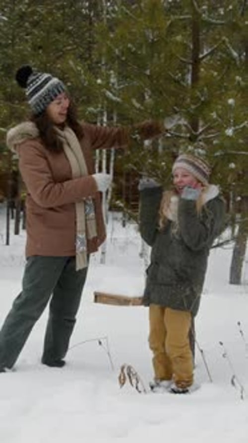 Vertical of Mother Giving Young Daughter Snow Shower in Winter Forest