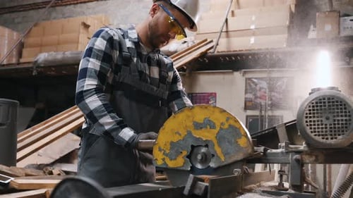 Male factory worker in hard hat and uniform is cutting metal by a big saw