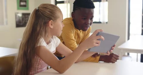 School children holding gray tablet in classroom, girl angling while boy leaning in, seeing screen