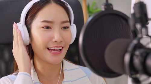 Asian podcaster in headphones using microphone broadcasting in studio.