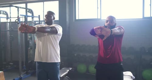 Two African-American men in tees clasping hands swinging arms stretching shoulders by weight rack