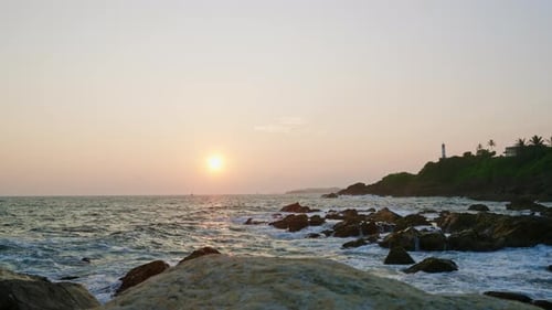Lighthouse Stands on Hill at Sunset Waves Crash Against Rocks Below Golden Hour Light Reflects on