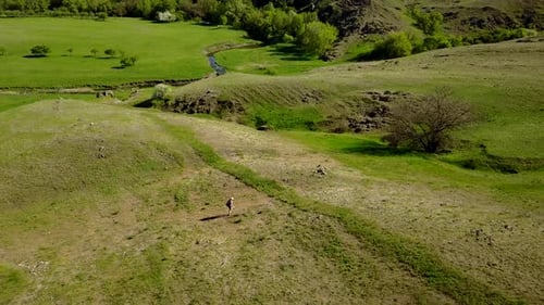 Aerial Video of Man Walking in Picturesque Valley on Green Grass Towards a River Drone Tracking Shot
