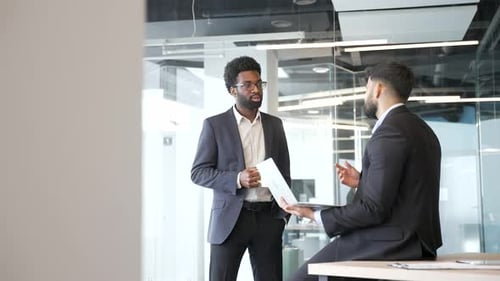 Two businessmen talking during break in modern office, holding coffee cup and using laptop.