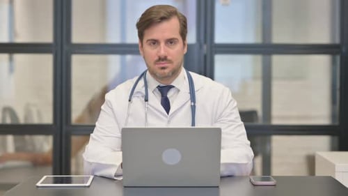 Arzt schaut in die Kamera, während er im Krankenhaus am Laptop arbeitet
