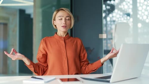 Young woman is meditating with her eyes closed while sitting at a workplace in a office.