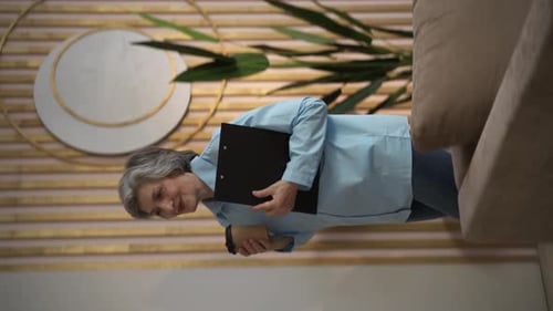 Vertical Video An Elderly Woman Walks Around the Office Holding a Tablet in Her Hands A Green Plant