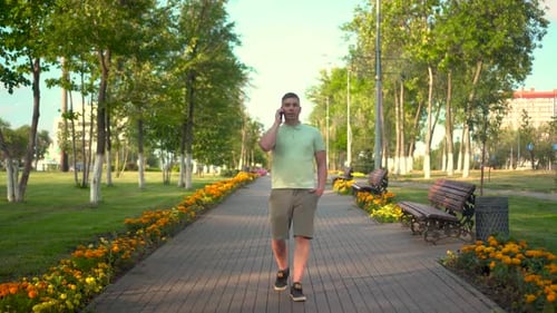 Un jeune homme marche dans le parc et parle au téléphone Un homme en polo et short marche le long d'un
