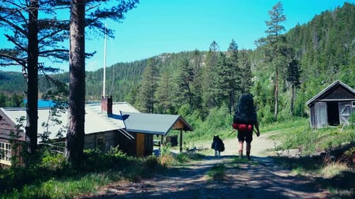 Backpacker With His Dog Passing On The Road Through Rural Village Near Forest Mountain.