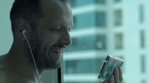 Cheerful young man enjoys a movie on his smartphone at home by the window