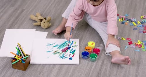 Fille asiatique d'âge préscolaire peignant de manière colorée avec un pinceau sur du papier alors qu'elle est assise sur le sol de la maison à côté