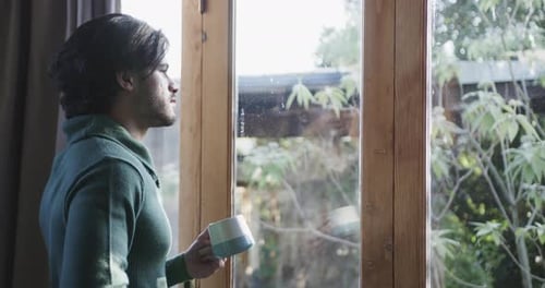 Thoughtful caucasian man with cup of coffee and looking out window at home, copy space, slow motion