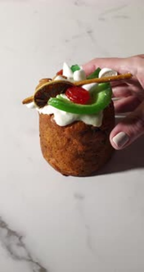 Close-up of a hand grabbing a festive cupcake topped with whipped cream, candied fruits, dried lime,