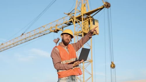 Construction Worker Directing Crane Operation with Laptop