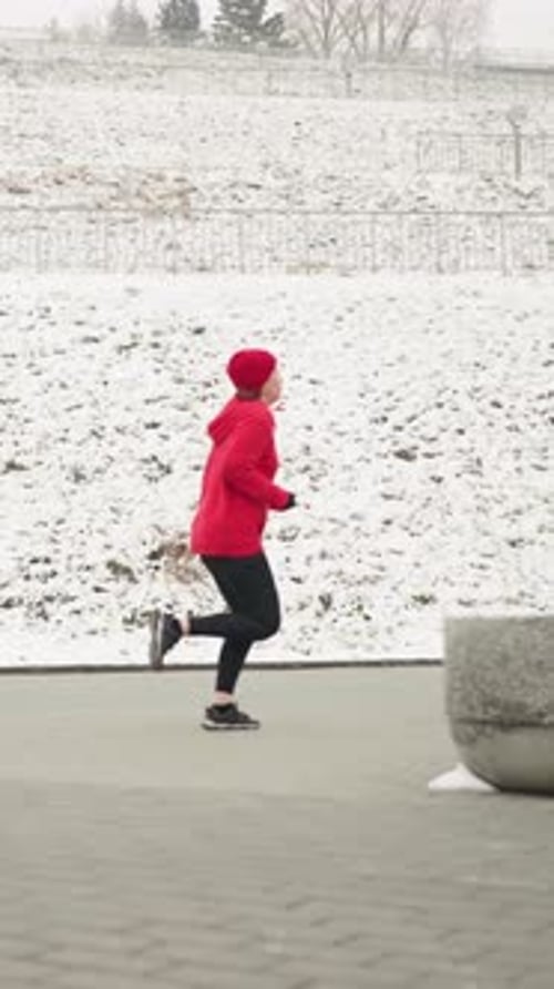 Side View Adult Jogging Outdoors Near Snowy Hillside in Winter with Red Hoodie