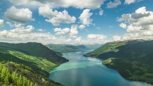 Timelapse of fluffy white clouds flowing over huge lake in middle of green mountains on a bright sum