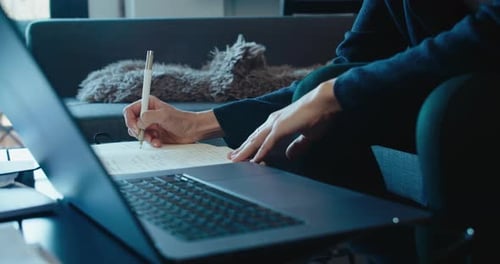 Adult Writing in Notebook with Laptop in Home