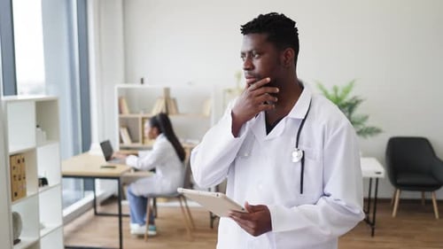 Doctor in Modern Office Using Tablet in Thoughtful Pose Near Colleague