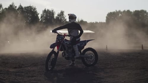 Sportsman in Uniform and Helmet Sits on Motorcycle in Dusty Field