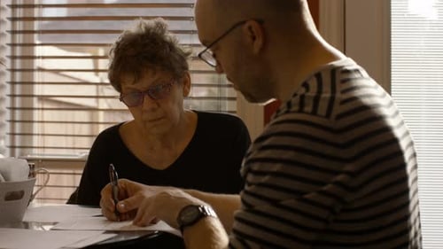 Senior Woman Signing Documents With Help From Caregiver Son At Home On Spring Afternoon Medium Shot
