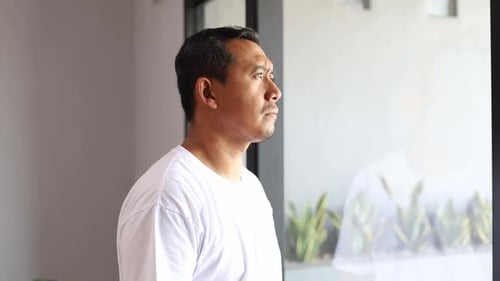 Trauriger, denkender asiatischer Mann am Fenster zu Hause aus Gründen der psychischen Gesundheit