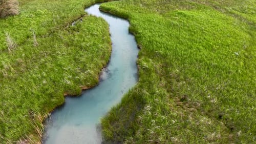 Aerial drone view of a winding blue stream flowing through vibrant green grassy marshland.