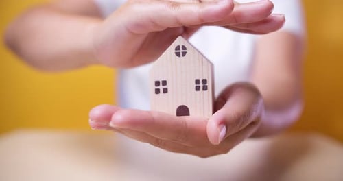 Hand of a woman holding and covering a wooden model house. Real estate home mortgage or buying and i