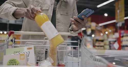 Closeup of Male Hand Shopping in Supermarket Putting Food in Cart and Using Smartphone