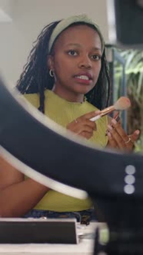Vídeo vertical: Escogiendo un pincel, una mujer con una banda verde aplicando rubor en el escritorio, filmando con una luz anular
