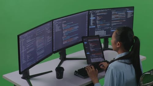 Seitenansicht einer asiatischen Frau, die mit einem Tablet arbeitet, während sie Code mit mehreren Computern auf einem grünen Bildschirm schreibt
