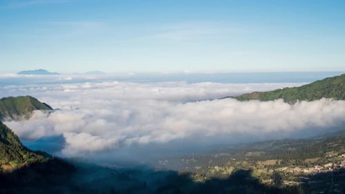 Fluffy moving clouds viewed from above, over a town ni a mountain valley with setting shade. Bromo,