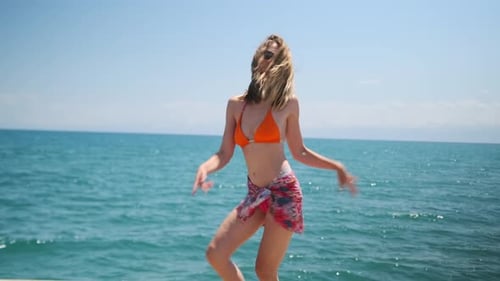 young woman in sunglasses dancing on wooden pier on turquoise water and blue sky background.