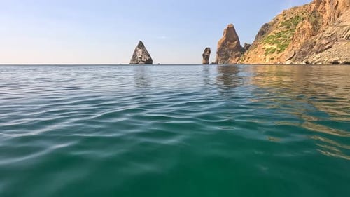 Sea Water Surface Camera Flies Over the Calm Azure Sea with Volcanic Rocky Shores on Background