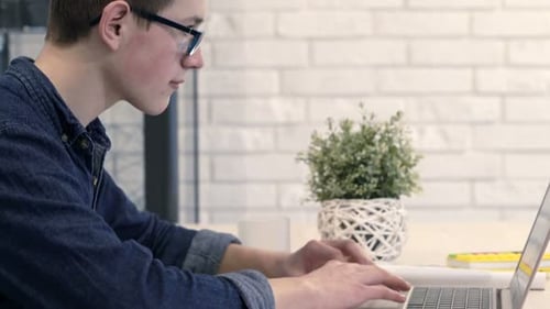 Junge mit Laptop und Notizbuch macht Hausaufgaben, sitzt am Tisch