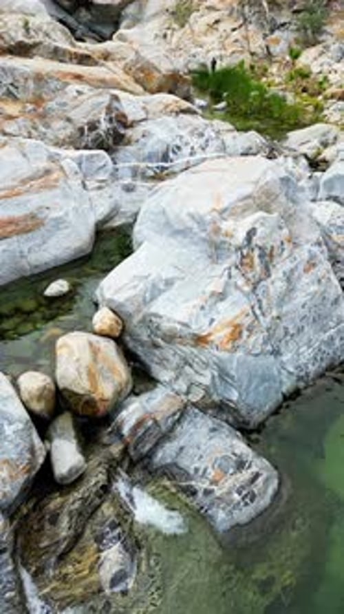 Aerial drone view of a calm natural pool surrounded by large gray rocks and crystal clear water. Ver