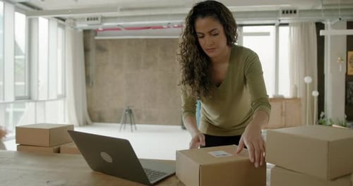 Woman Working on Laptop at Table in Office, Preparing E-commerce Parcels for Shipment