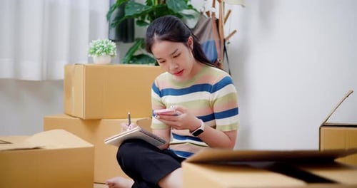 Woman Sits Among Boxes Using Phone and Notepad