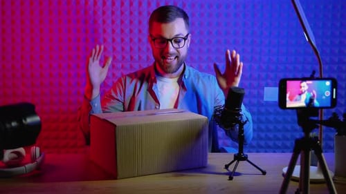 Young Smiling Blogger Unpacking Box with Present Sitting at Home Studio