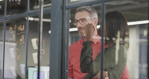 Focused diverse male and female colleague brainstorming writing on glass wall in office, slow motion